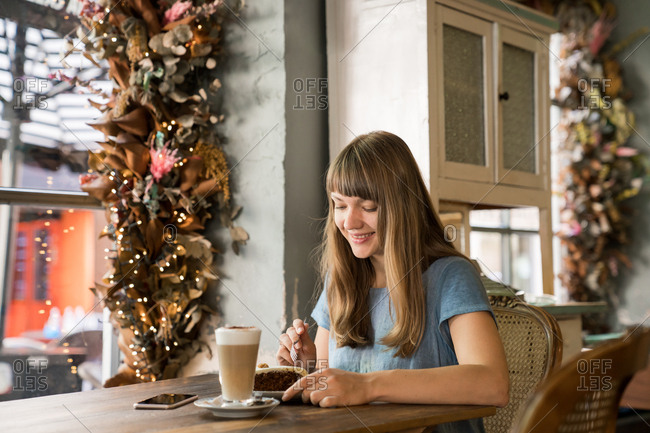 Blonde happy young female with bangs in casual blue T-shirt smiling and using a phone while holding cup of coffee in cozy cafe