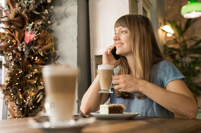 Blonde happy young female with bangs in casual blue T-shirt smiling and talking on smartphone while holding cup of coffee in cozy cafe