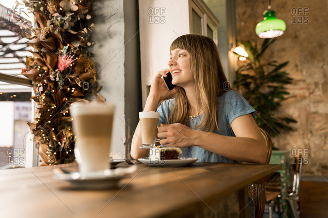 Blonde happy young female with bangs in casual blue T-shirt smiling and talking on smartphone while holding cup of coffee in cozy cafe