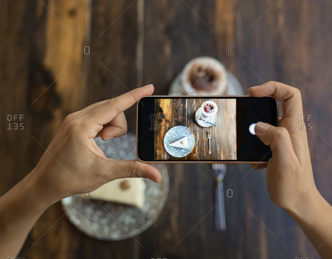 Unrecognizable woman taking photo by smartphone of piece of cake on plate with ornament and served coffee on wooden table