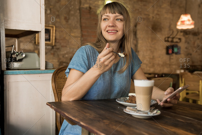 Blonde cheerful happy female with bangs in blue casual T-shirt holding spoon with piece of cake and sitting at table with coffee and dessert