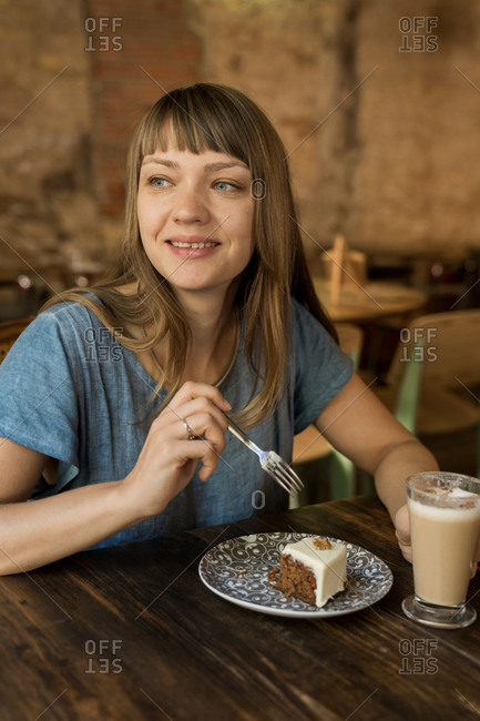 Blonde cheerful happy female with bangs in blue casual T-shirt holding spoon with piece of cake and sitting at table with coffee and dessert