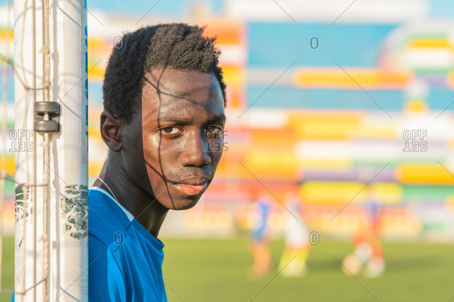 Ethnic teenager looking at camera while leaning on net on blurred background of football field on sunny day
