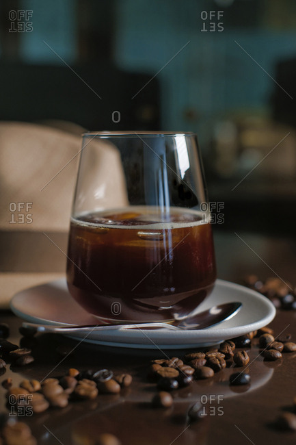 Delicious aromatic brown beverage in glass on round saucer standing among roasted coffee beans beside hat with brim on wooden glossy table