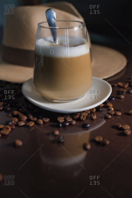 Delicious aromatic brown beverage with white foam in glass on round saucer standing among roasted coffee beans beside hat with brim on wooden glossy table