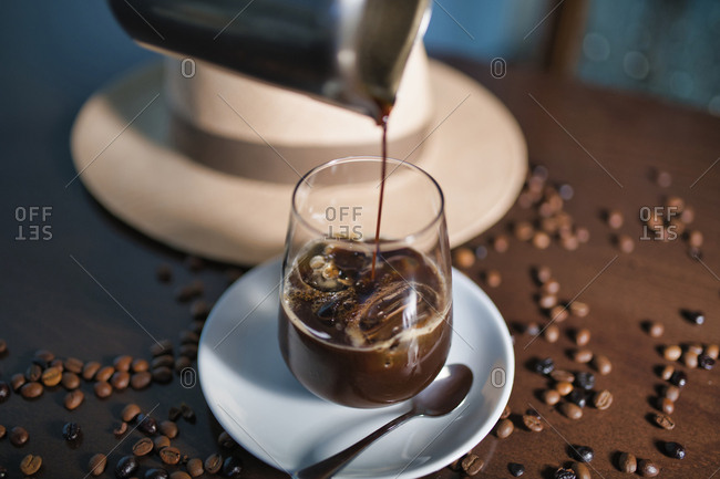 Delicious aromatic brown beverage in glass on round saucer standing among roasted coffee beans beside hat with brim on wooden glossy table