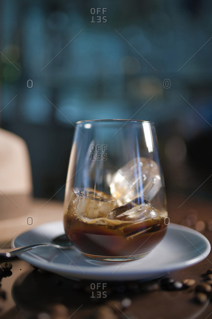 Delicious aromatic brown beverage in glass on round saucer standing among roasted coffee beans beside hat with brim on wooden glossy table