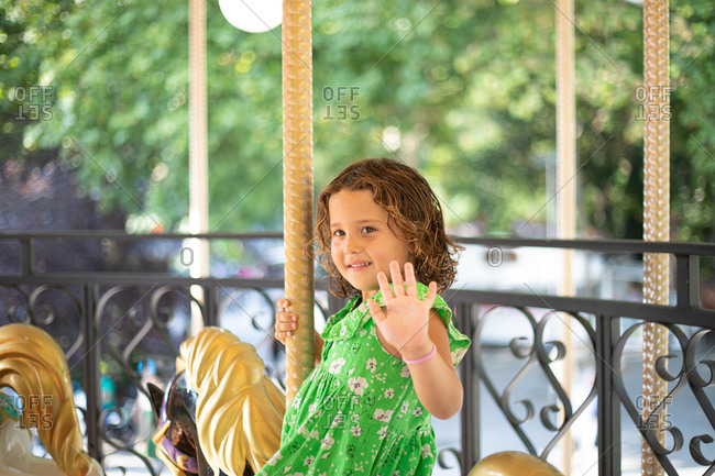 Smiling pensive child in green casual clothes looking away and waving hand while riding fabulous carnival pony in amusement park