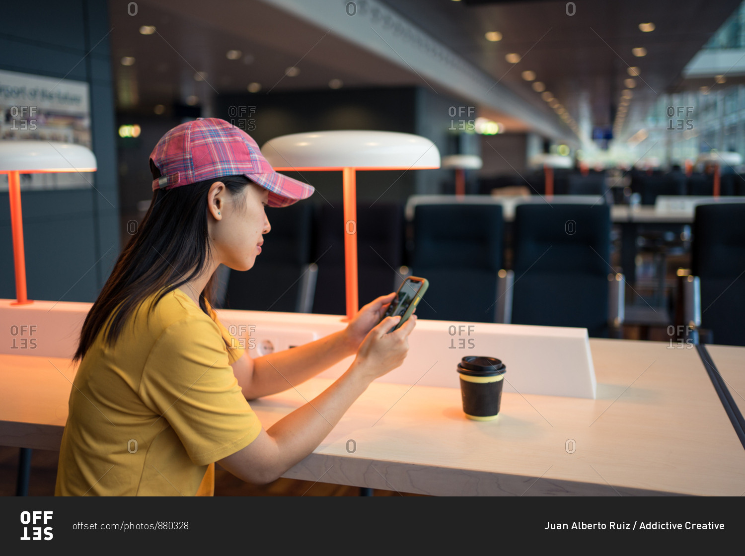 Side view of Asian woman in cap surfing mobile phone and drinking coffee from disposable cap at table before schedule board in airport