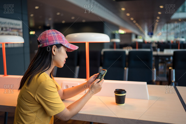 Side view of Asian woman in cap surfing mobile phone and drinking coffee from disposable cap at table before schedule board in airport