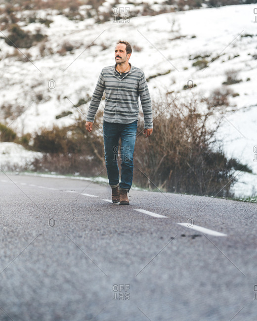 Adult male in stripped jumper and jeans walking on country roadway with hills covered by snow on background on gloomy cloudy day