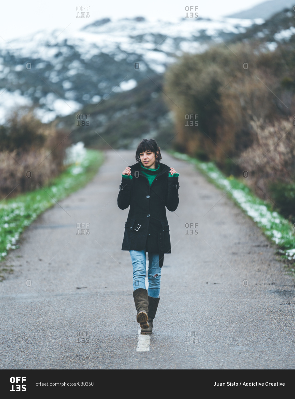 Pretty young female in black coat and jeans walking on empty country road with hills covered by snow on background on gloomy cloudy day