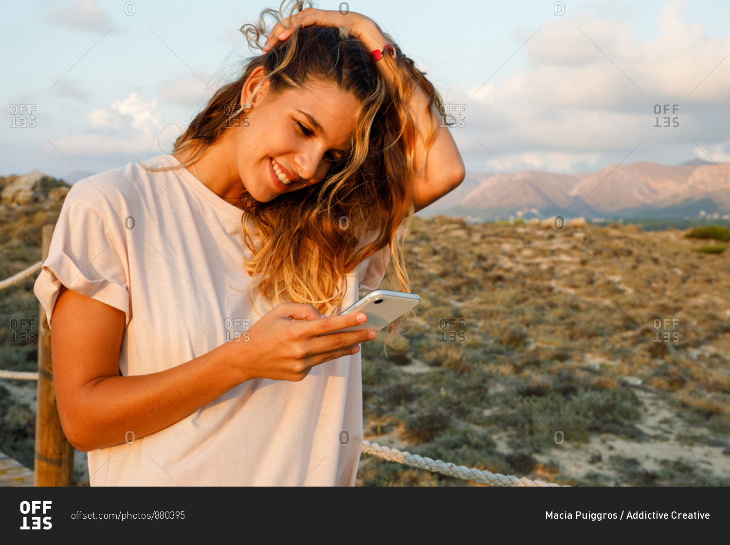 Satisfied curly haired female traveler in casual white shirt touching hair while web surfing on mobile phone with beautiful landscape on background