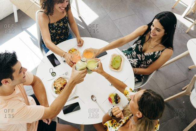 From above multi ethnic young people clinking glasses of fresh fruit beverages and proposing toast while sitting at table with healthy snacks in cafe