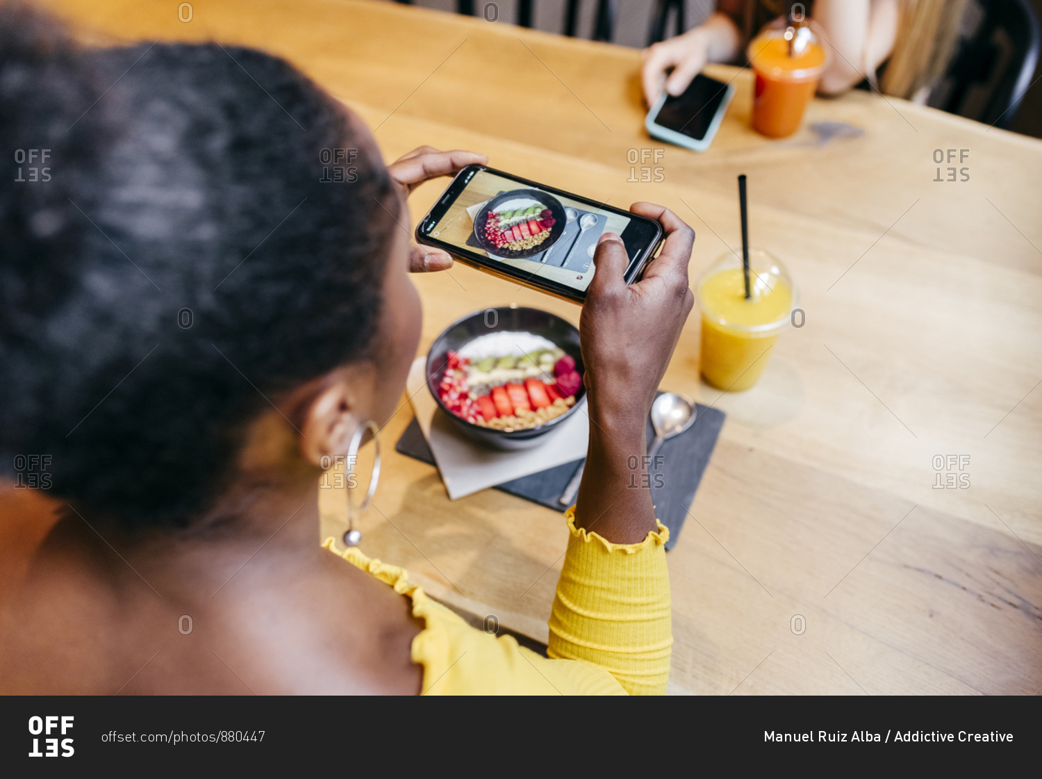 From above of young African American female sitting at wooden table with smoothie bowl and cup of fruit juice and taking picture of food with smartphone