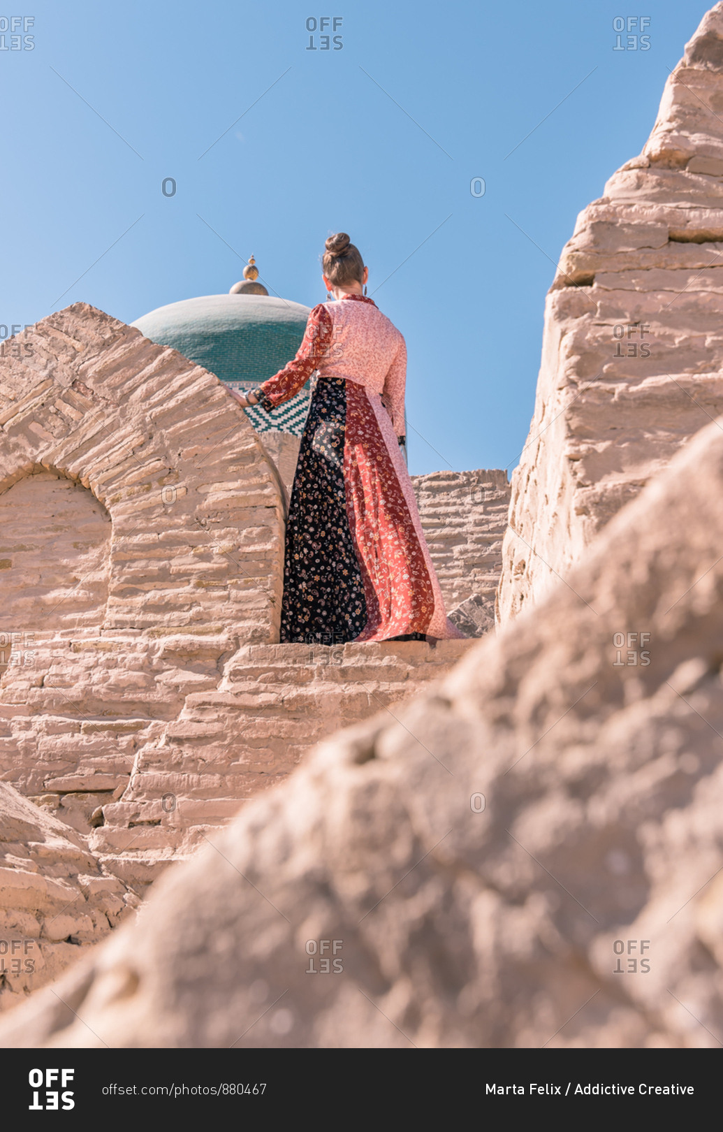From below back view of calm female in traditional long dress standing on stone stairs and looking away against old blue dome of mausoleum of Pahlavan Mahmud in Khiva