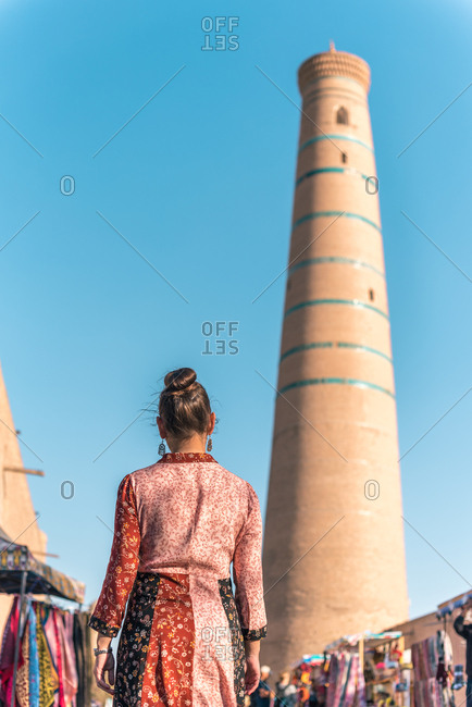Back view of female in colorful dress walking along open air oriental market with traditional clothes and souvenirs near tall brown brick tower of Islam Hodja minaret in Khiva under cloudless blue sky