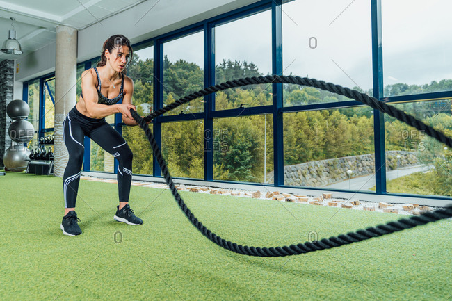 Full length focused strong woman in sportswear doing exercise with battle ropes during training in modern gym