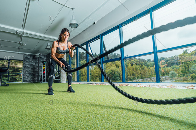 Full length focused strong woman in sportswear doing exercise with battle ropes during training in modern gym