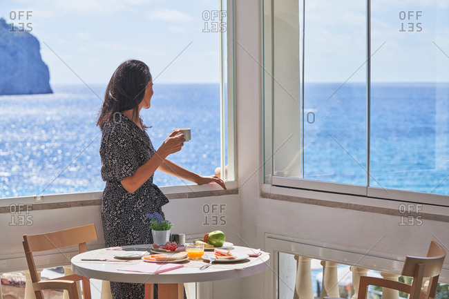 Side view of thoughtful woman drinking coffee standing nearby window enjoying shiny calm water in light cozy terrace with served for breakfast table in Mallorca, Spain