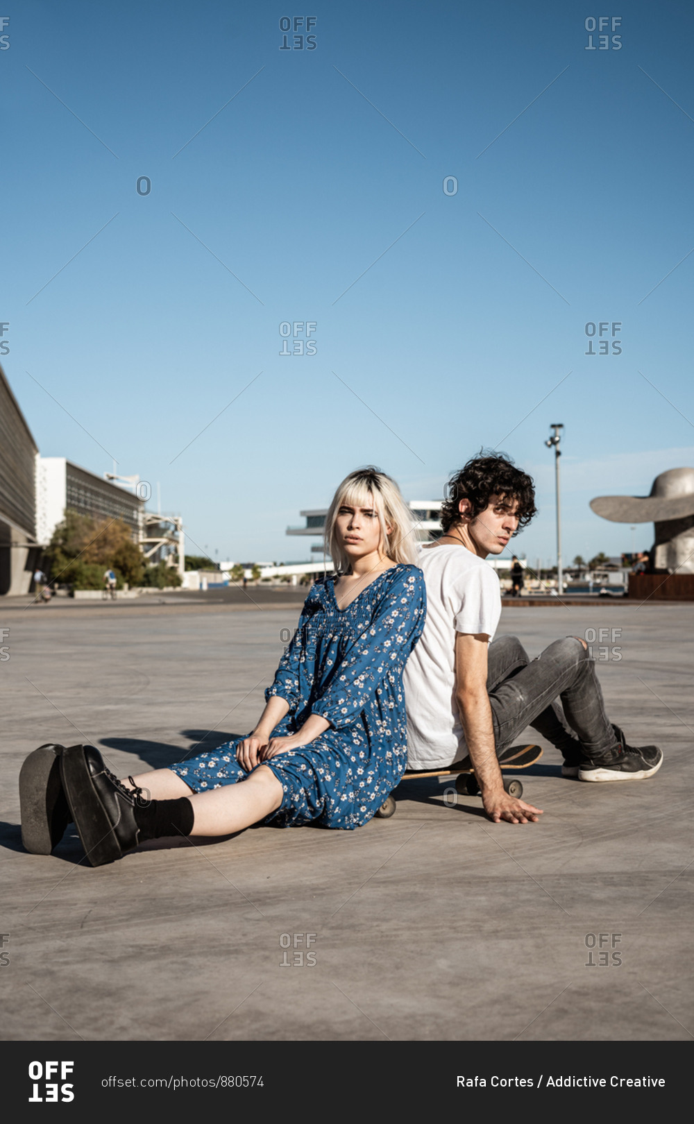 Attractive sensual blond woman looking at camera sitting with boyfriend on skateboard and dreaming against blue sky and blurred modern buildings on square
