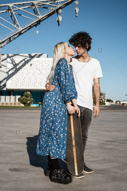 Young trendy loving couple standing and leaning on skateboard on square against blue sky and blurred modern buildings in windy weather