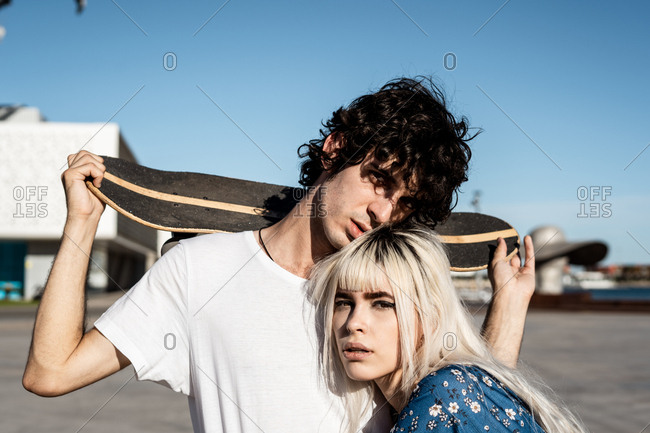 Attractive sensual blond woman with black eyebrows looking at camera with curiosity while embracing dark haired young man in white shirt