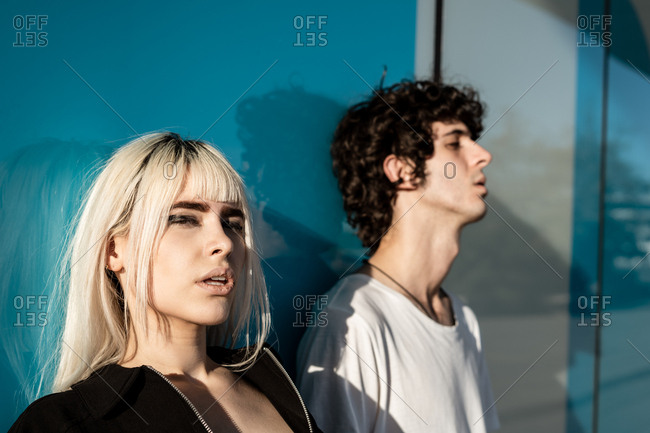 Magnificent blond provocative woman in black clothes standing beside tall man with closed eyes while standing against blue wall with reflection of street in sunlight