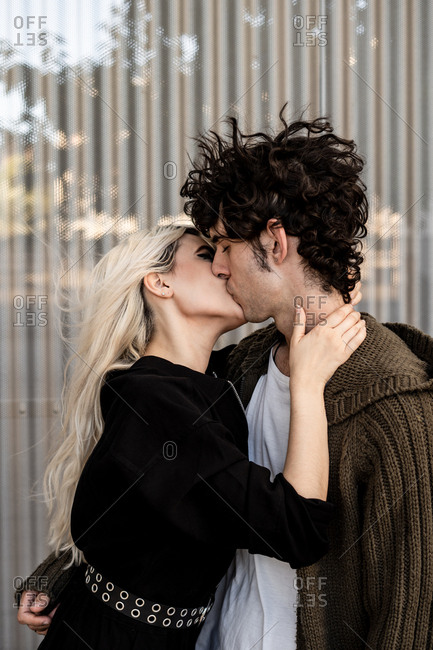 Side view of adult blond woman and young curly dark haired man kissing on street against striped wall of modern building in windy weather