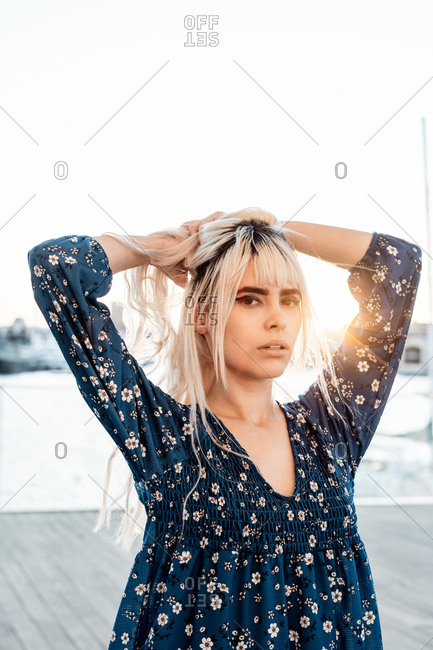 Gorgeous pensive blond female with black eyebrows in blue oversize dress looking at camera with curiosity while standing and touching hair on street during sunset