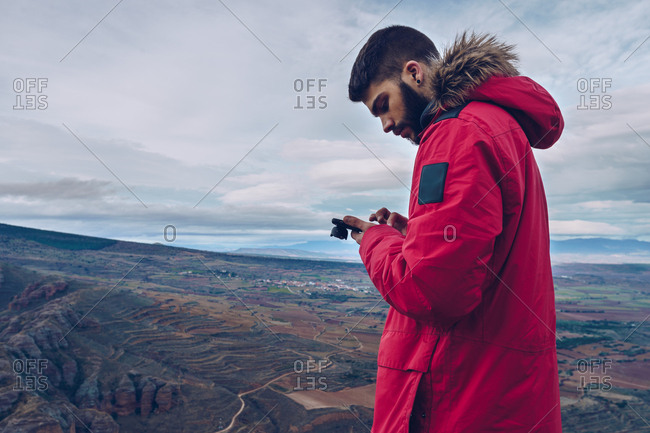 Side view of attentive man in jacket using smartphone while standing at cliff over breathtaking view