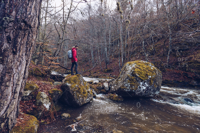Side view of bearded man with backpack walking on rocks by mountain river in cold leafless trees on autumn day