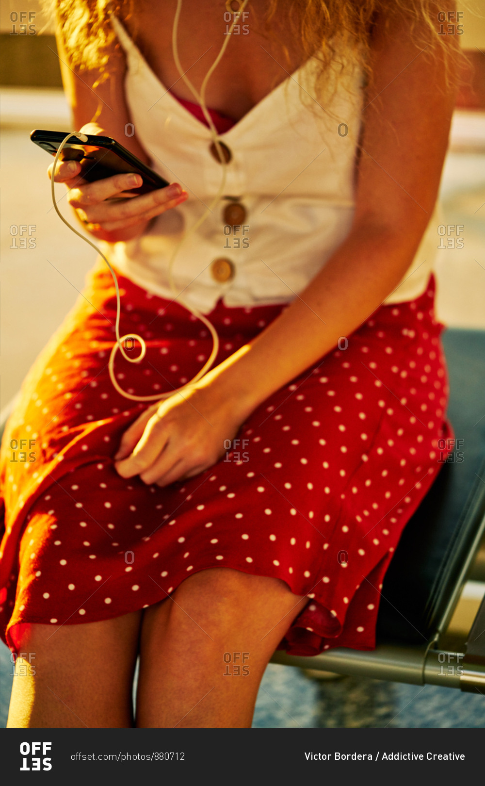 crop woman in earphones listening to music with mobile phone while chilling on metal bench in airport of Texas