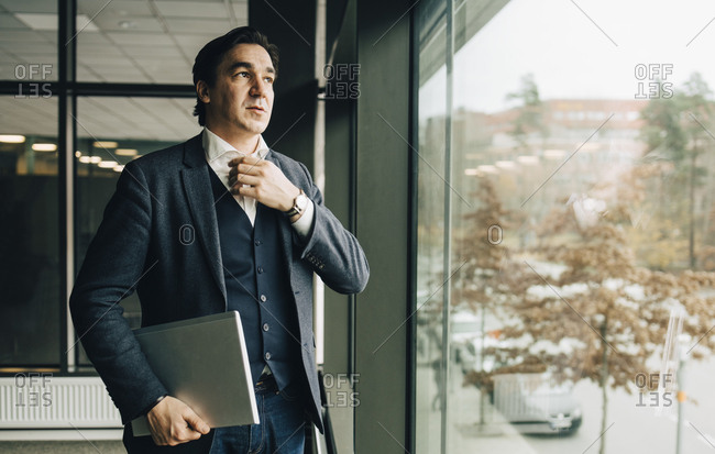 Businessman adjusting collar while standing with laptop by window in office