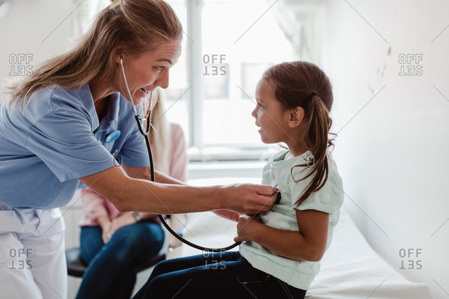 Female pediatrician examining girl's heartbeat through stethoscope while mother sitting in background at clinic
