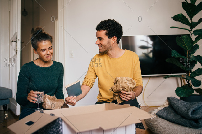 Smiling boyfriend showing novel to girlfriend while removing glasses from box in living room at new home