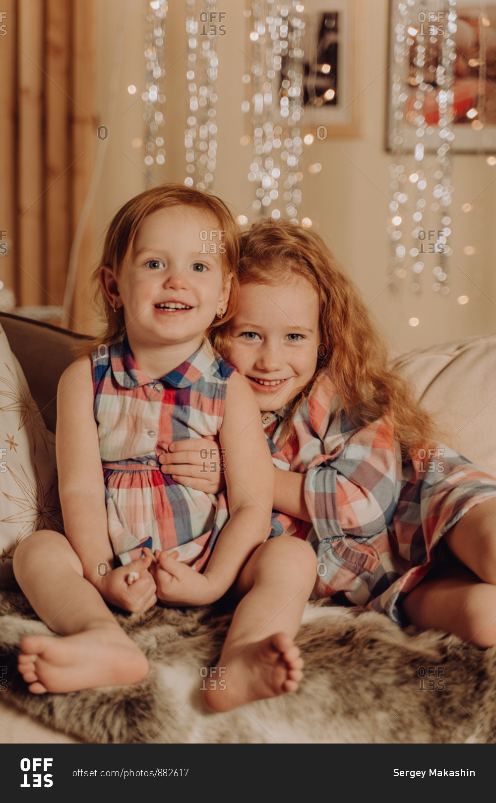 Two young sisters with red hair hugging and smiling with the holiday lights in the background