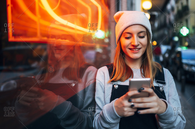 Smiling young woman standing next to neon light using her smartphone at night