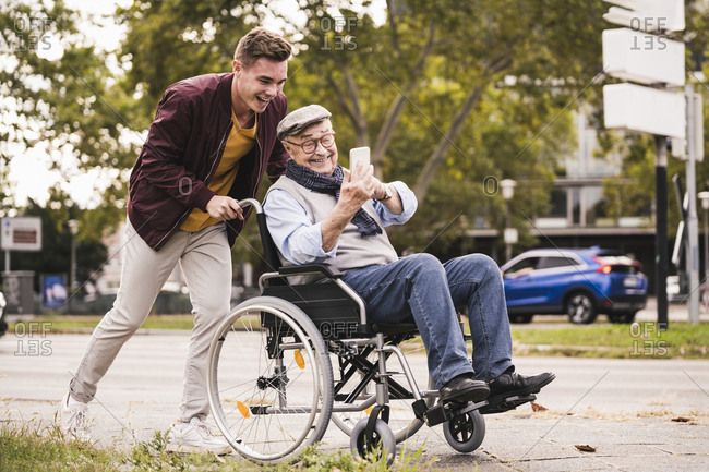 Smiling young man pushing happy senior man with smartphone in wheelchair