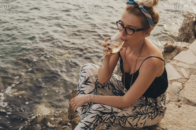 Pretty stylish woman sitting on seacoast and enjoying sunset drinking wine.