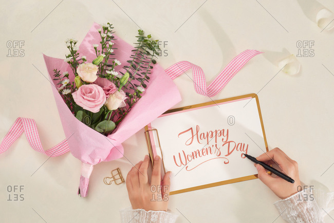 Woman hands writing Happy Valentines Card on paper over white background with flower bouquet and ribbon. Top view, flatlay.