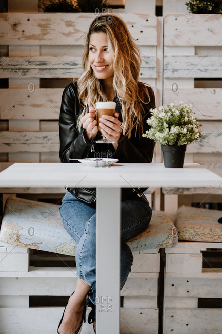 Long haired trendy beautiful blonde woman drinking from a glass of delicious foamy coffee while looking away