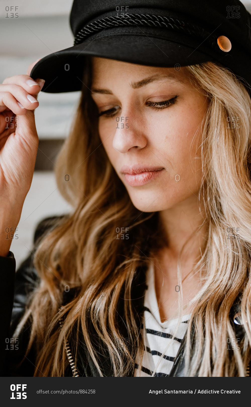 Stylish beautiful confident woman in leather jacket looking away while touching trendy black cap in her head on wooden wall