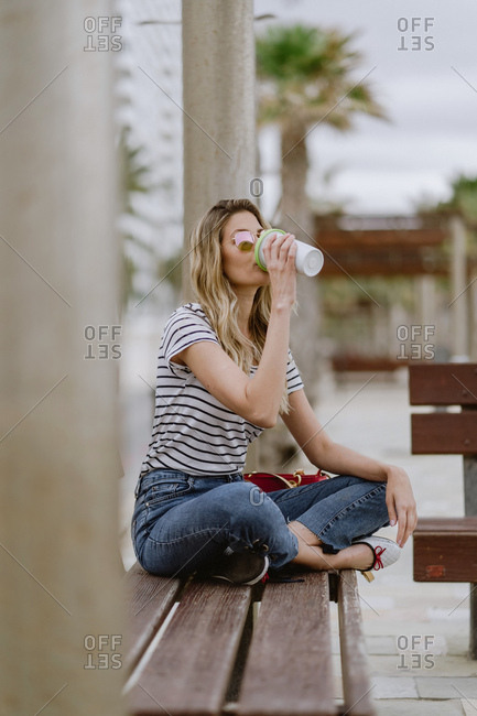Side view of cheerful casual young female drinking from takeaway cup of coffee sitting on city bench at seafront on summer day
