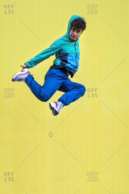 Cheerful curly ethnic male dancer in active blue wear jumping with crossed legs and covering face with hand on yellow background
