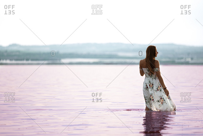 Back view of romantic female in flowing gentle dress walking in shallow water of exotic pink lake at Torrevieja at Spain