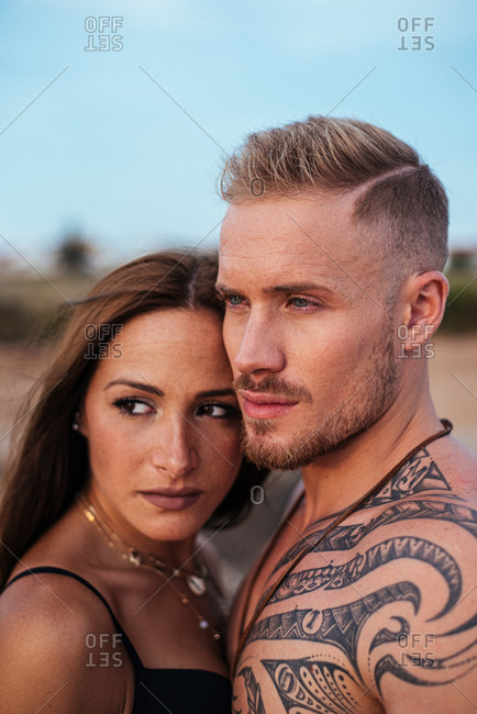 Brutal shirtless tattooed male and sensual woman in swimsuit standing close and looking away with blue sky on background at Spain