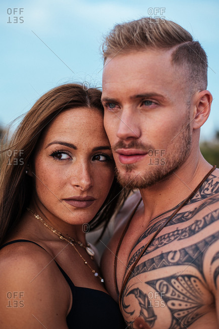 Brutal shirtless tattooed male and sensual woman in swimsuit standing close and looking away with blue sky on background at Spain