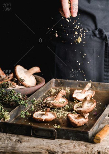 Crop person sprinkling with herbs fresh brown mushrooms Shiitake on metal tray at rustic wooden table