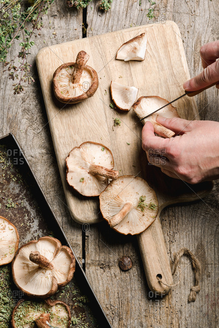 From above crop cook cutting caps of fresh brown Shiitake mushrooms on wooden cutting board at shabby rustic table